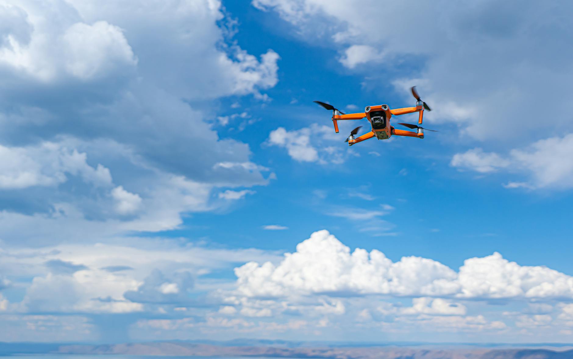 An orange drone soaring in a clear blue sky with fluffy clouds, capturing aerial views.